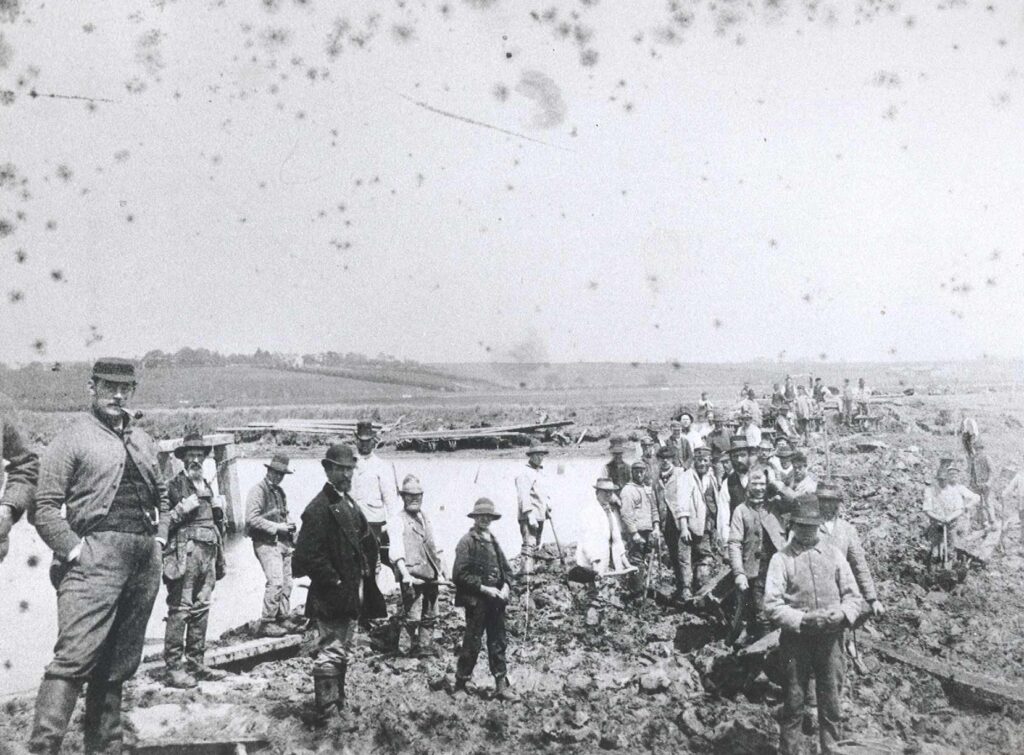 A black and white photograph of men in a line. Behind them is a large body of water and muddy fields.