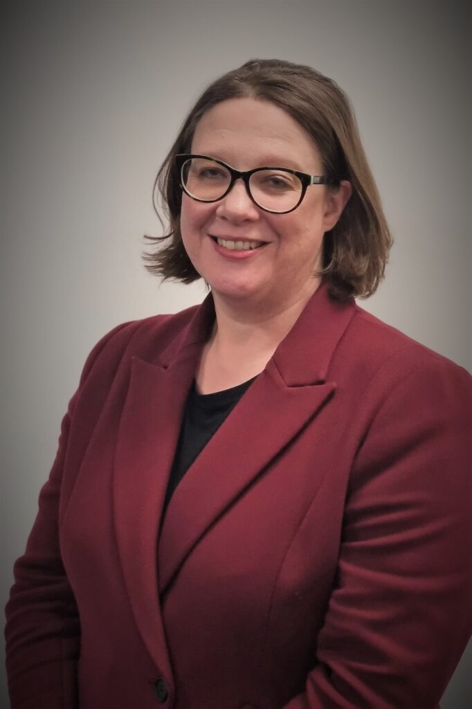 A white woman with glasses smiling. She is wearing a red suit.