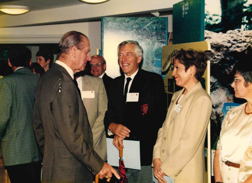 A coloured photograph of an event. A white man, Prince Phillip, is talking to two white people - a man and Annette Whybrow, who is smiling at the Prince.