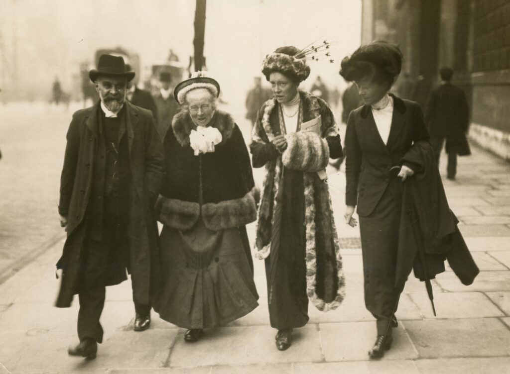 A black and white photograph of four people, in conversation, walking down a street.

From left to right:
There is a man in a black hat, with a large beard and moustache. Next to him is an older woman. Two younger women, one in a fur coat, walk to the right.