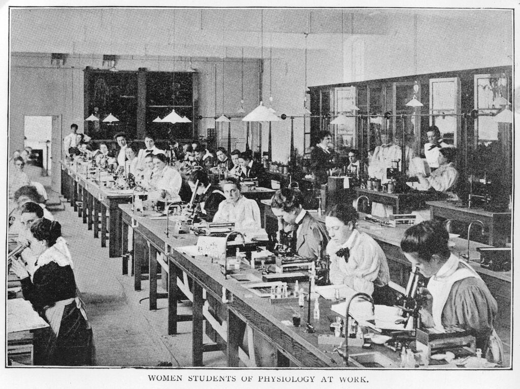 A black and white photograph of a science laboratory. 

Women sit on a long row of tables, with science equipment in front of them. Some look at the camera, whilst others are working.