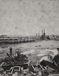Two women sitting by old machinery, with muddy construction site in the background.