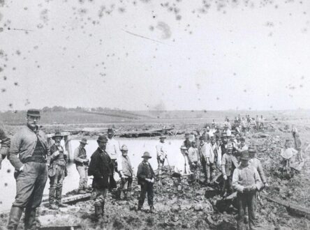 A black and white photograph of men in a line. Behind them is a large body of water and muddy fields.