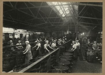 A black and white photograph of women, working with large barrels, in a factory.
