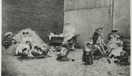 A black and white photograph of children and nursemaids in a small factory yard.
