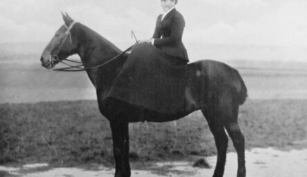 A black and white photograph of a woman riding a black horse.