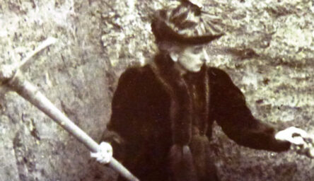 A black and white photograph of a finely dressed woman with a pickaxe in a dig.