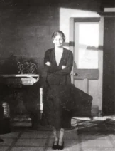 A black and white photograph of a woman standing in a house with her arms crossed.