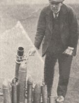 A black and white photograph of a men stood next to munitions.