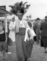 A black and whilte photo of a Black man wears clothing which looks flamboyant and reminisent of traditional African ceremonial clothing. He wears a headdress with feathers. He is pointing upwards with his right hand. The man is tall and in the background are white people dressed smartly at a horseracing event.