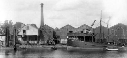 A black and white photograph of a large ship in a dock, with a factory in the background.