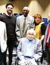 A group of men and a woman of varying ages stand next to each other. One man sits in a chair at the front of the group.