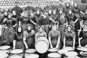 An old black and white big group of fisher men and women sit and stand in three rows facing the camera. A women in the front row sits with a large barrell in front of her. over a large wooden creates to process the fish.