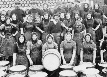 An old black and white big group of fisher men and women sit and stand in three rows facing the camera. A women in the front row sits with a large barrell in front of her. over a large wooden creates to process the fish.