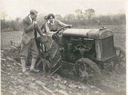 Black and white photo of a woman on a tractor in a field with a man standing next to her leaning on the vehicle.