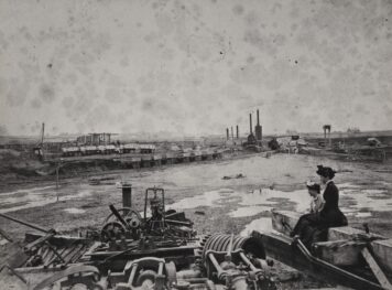 A black and white photograph of women sitting beside old machinery at a dock.