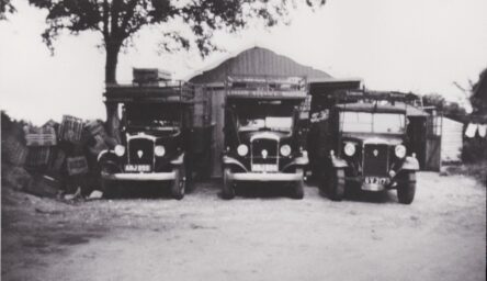 black and white photo of three old trucks lined up in ront of a barn.