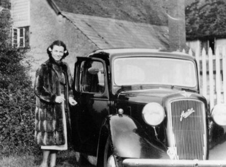 Black and white photo of a woman in a long fur coat, standing next to an old 1930s car.