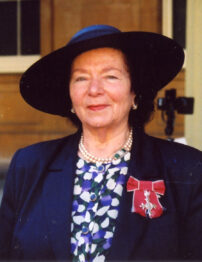 Head and shoulders shot of a white woman wearing a smart blue jacket and dress, with a dark blue het and a large red medal on her lapel.