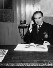 Black and white photo of a white man sitting at a desk, looking at a newspaper, dressed in a suit. He's talking on the phone.