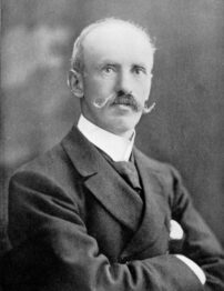 A black and white photograph of a light-skinned man with a large moustache posing, with a faded black background.