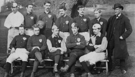 A black and white photograph of 12 men, some sitting on a bench and others standing behind them, dressed in an old football kit. There is a large tree and fields in the background.