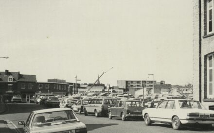 A black and white image of old cars parked along the street, a car park and buildings in the background.