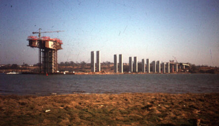 A photograph of a bridge being built, with a large crane. There is grass in the foreground, and the construction is taking place across a river.