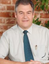 A photograph of a white man. Behind him is a brick wall and a green plant.