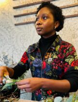 A black woman with short black hair arranges a bouquet of flowers. She wears a colourful jumpsuit,
