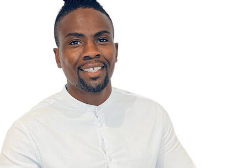 A Black man is against a white background. His braids are in a top knot and he wears a plain white top.