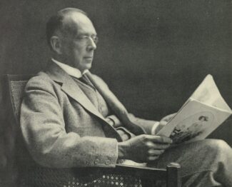 A black and white photograph of a man in spectacles sitting in a chair reading a magasine.