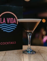 An espresso martini is in a shallow cocktail glass in front of a colourful menu.
