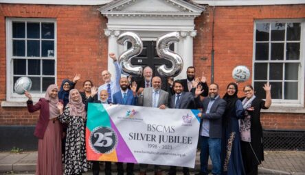 A group of people outside a building, holding a banner. Some are holding celebration balloons.