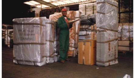 A black man standing in a warehouse, in a hard hat holding a clipboard. There is packaged goods around him.