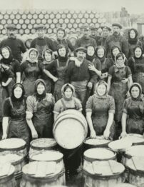 A black and white photograph of women in headscarves standing in front of barrels.