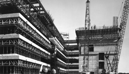 Black and white photograph of two men standing on a construction site of a large building.