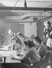 A black and white photograph of women sitting around a table, one using a telephone.