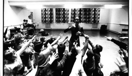 A black and white photo of children in a dance class. They are reaching into the air towards their female dance instructor.