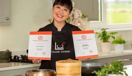 A woman of South east Asian origin standing in a kitchen. In her hands are two award certificates.