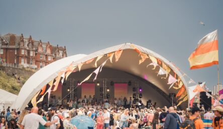 A large festival stage with a white tent. Colourful bunting blows in the wind. A large crowd stands in front of the tent. facing the stage.