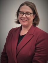 A white woman with glasses smiling. She is wearing a red suit.