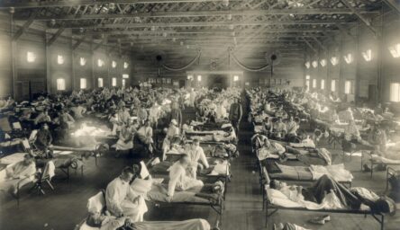 A black and white photograph of rows of make-shift hospital beds in a large room. The beds are busy with patients, and doctors attend them wearing masks.