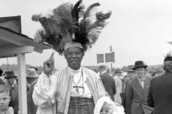 A black and white photograph of a black man looking at the camera, smiling. He wears an embroidered waist coat, and has a large feather headress on.