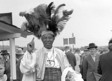 A black and white photograph of a black man looking at the camera, smiling. He wears an embroidered waist coat, and has a large feather headress on.